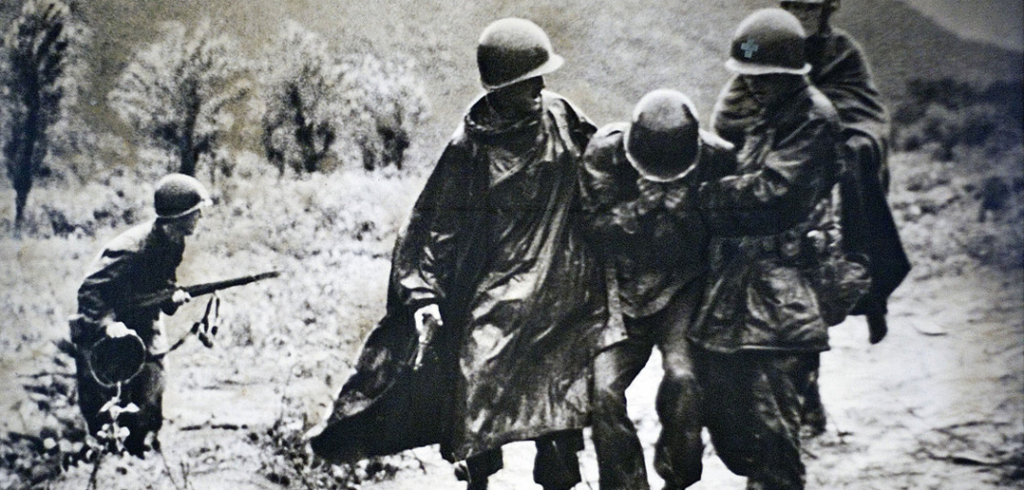 During the Korean War, a wounded soldier is assisted by posthumous Medal of Honor Recipient (and candidate for sainthood) Army chaplain Father Emil Kapaun. 